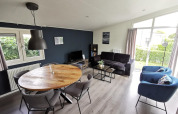 Modernes Wohnzimmer in der „Comfort“-Hütte im De Woudhoeve, Niederlande, mit Sofa, Esstisch und großen Fenstern.