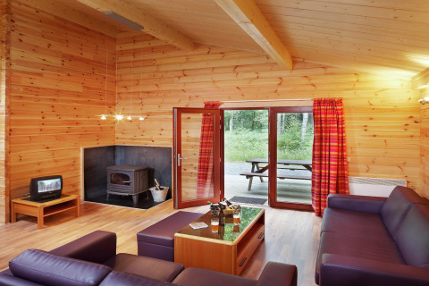 Salon du chalet en bois 'De Boshut' en Belgique avec canapés, poêle à bois et vue sur la forêt.