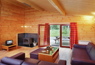 Salon du chalet en bois 'De Boshut' en Belgique avec canapés, poêle à bois et vue sur la forêt.