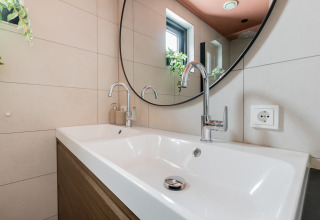 Lavabo moderno con due rubinetti, grande specchio rotondo e finestra presso la Forest villa «De Bosburcht» nei Paesi Bassi.