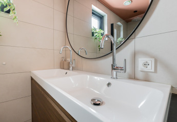 Modern bathroom sink with two faucets, large round mirror, and window at Forest villa «De Bosburcht» in the Netherlands.