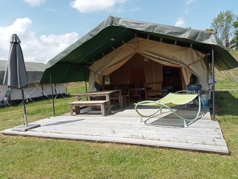 Safari tent with a covered wooden deck, picnic table, umbrella, and lounge chair in a grassy area.