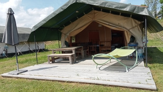 Safari tent with a covered wooden deck, picnic table, umbrella, and lounge chair in a grassy area.