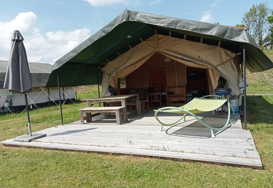 Safari tent with a covered wooden deck, picnic table, umbrella, and lounge chair in a grassy area.