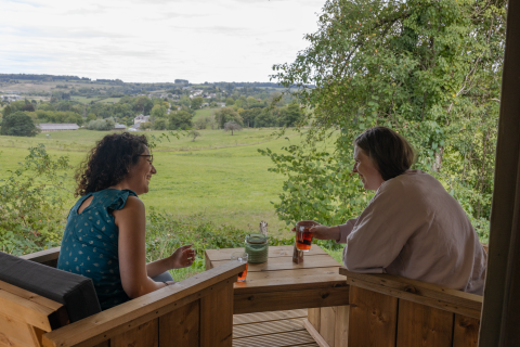 Dos mujeres disfrutan bebidas y conversan en la terraza de Stardust 40 Family Lodge, con vistas a los campos verdes.