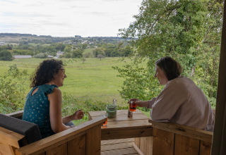 Dos mujeres disfrutan bebidas y conversan en la terraza de Stardust 40 Family Lodge, con vistas a los campos verdes.