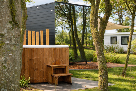 Udendørs jacuzzi af træ ved River Mirrorlodge i Holiday park Mölke, omgivet af grønne træer og hytter.