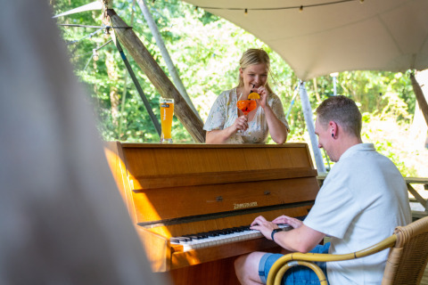 Two people enjoy music and drinks by a piano outdoors at Gooise Heide holiday park in North-Holland.