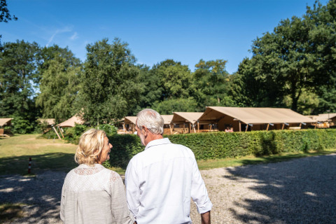 Couple senior se promenant au parc de vacances Gooise Heide en Hollande-Septentrionale, avec tentes de glamping en arrière-plan.