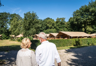 Senior couple strolls at Gooise Heide holiday park in North Holland, Netherlands, with glamping tents and greenery visible.
