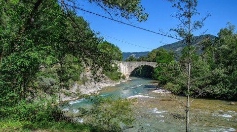 Stenbro henover en flod omgivet af tæt skov ved Flower Camping la Pinède i Auvergne-Rhône-Alpes, Frankrig.