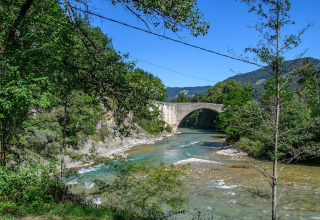Puente de piedra sobre un río claro, rodeado de bosque en Flower Camping la Pinède, Auvergne-Rhône-Alpes, Francia.