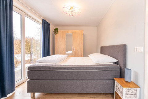 Camera luminosa con letto grigio, grande finestra, tende blu e guardaroba in legno a Villa BushBush, De Kremmer, Paesi Bassi.