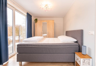 Camera luminosa con letto grigio, grande finestra, tende blu e guardaroba in legno a Villa BushBush, De Kremmer, Paesi Bassi.