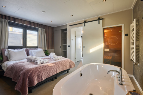 Chambre du Happy Oak wellnesslodge avec lit double, baignoire indépendante et porte menant à un sauna.