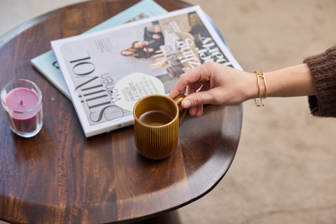 Een hand neemt een koffiekopje op een tafel met tijdschriften en een kaars in Happy Oak wellnesslodge.