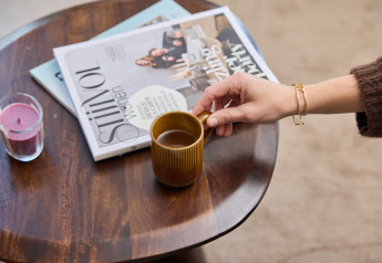 Een hand pakt een koffiekopje op een tafel met tijdschriften en een kaars bij Happy Oak wellnesslodge.