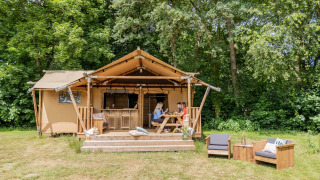 Tente safari Dreamer 40 en forêt, personnes à une table sur terrasse, fauteuils en bois sur la pelouse.