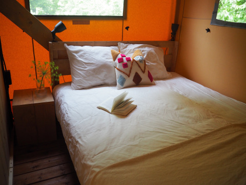 Gemütliches Bett mit weißer Bettwäsche, bunter Kissen und Buch in einem Safari-Zelt in Arden Parks, Frankreich.