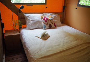 Knus bed met witte lakens, kleurrijk kussen en open boek in een safaritent in Arden Parks, Frankrijk.