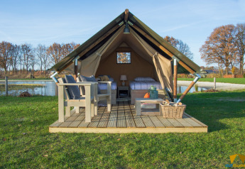 Safari tent with wooden deck, cozy outdoor furniture, and scenic lake view in a grassy landscape.