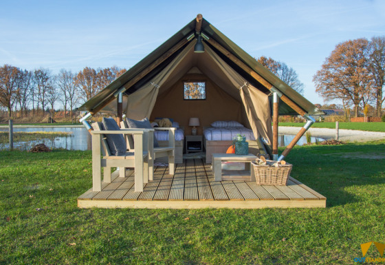 Tienda safari con terraza de madera, muebles al aire libre y vistas a un lago rodeado de naturaleza.