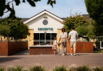 Eine Familie läuft Hand in Hand in Richtung der Rezeption einer gemütlichen Lodge mit Gartenanlage.