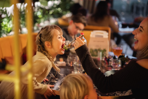 Een vrouw voert een vrolijk meisje in een sfeervolle lodge-restaurant met warme, gezellige inrichting.