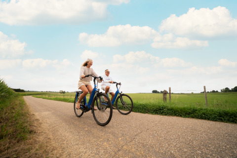 Twee personen fietsen op een rustige landweg met groene weiden vlakbij een lodge.