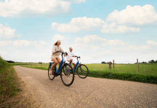 Deux personnes font du vélo sur une route de campagne près d’un lodge, entourées de champs verts.