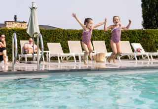 Twee kinderen springen samen in een zwembad bij een lodge terwijl volwassenen rusten op ligstoelen.