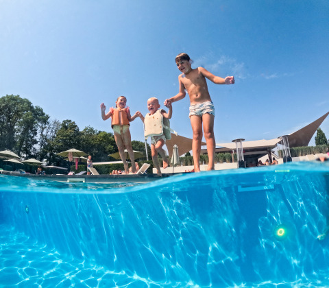 Kinderen met zwembandjes springen in het zwembad bij lodge Biebosch, Resort Mooi Bemelen in Nederland, op een zonnige dag.