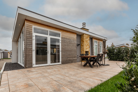 Lodge moderne avec vaste terrasse carrelée, table à manger extérieure, chaises et grandes baies vitrées.