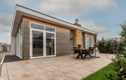Lodge moderne avec vaste terrasse carrelée, table à manger extérieure, chaises et grandes baies vitrées.