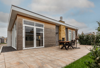 Moderne lodge met ruime betegelde patio, buitentafel, stoelen en grote ramen onder een bewolkte hemel.