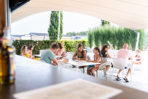 Des personnes se détendent et discutent sur la terrasse d’un lodge sous un grand auvent en été.