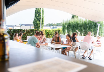 Des personnes se détendent et discutent sur la terrasse d’un lodge sous un grand auvent en été.