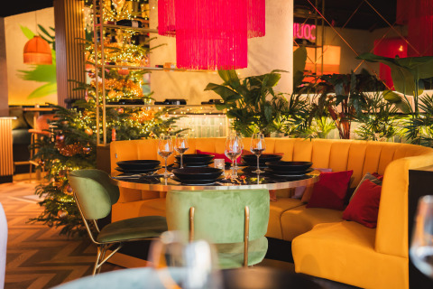 Salon de restaurant chic avec canapé jaune, chaises vertes, sapin de Noël et plantes luxuriantes.