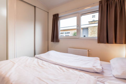 Slaapkamer in een lodge met tweepersoonsbed, witte lakens, grote ramen en bruine gordijnen.
