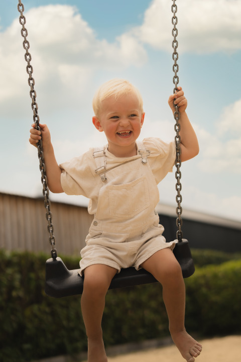 Young child in light overalls smiling on a swing outdoors at Biebosch, Resort Mooi Bemelen, Netherlands.