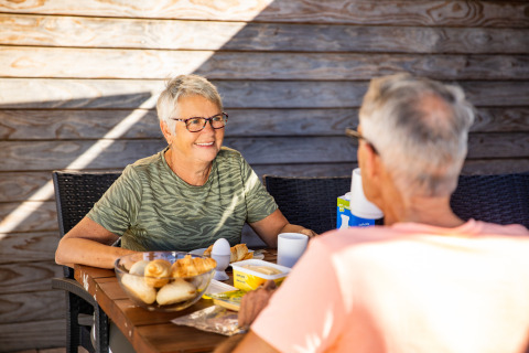 Deux personnes âgées prennent le petit-déjeuner ensemble à l’extérieur, assises à une table de lodge en bois.