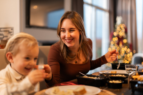 Une mère et son enfant partagent un repas chaleureux à table, avec un sapin de Noël illuminé derrière.