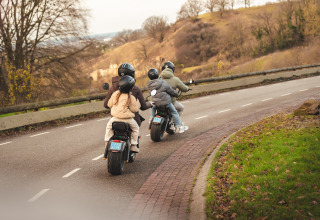 Deux motos transportant deux personnes chacune roulent sur une route sinueuse entourée d'arbres et de collines.