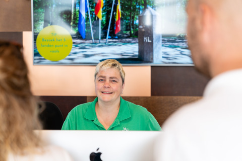 Réceptionniste accueillant des clients à l’accueil du Resort Mooi Bemelen aux Pays-Bas devant un panneau d'information.