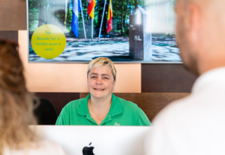 Réceptionniste accueillant des clients à l’accueil du Resort Mooi Bemelen aux Pays-Bas devant un panneau d'information.