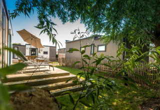 Terrasse med liggestole og parasol i ferieparken Holiday Village Knokke, omgivet af grønne træer og hytter.