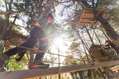 Children with harnesses balance on a wooden bridge at Spina Family Collection adventure park in Italy.