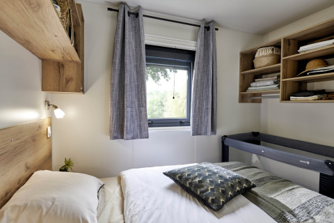 Bedroom in Mobile Home «New Horizon» at Flower Camping Provence Vallée, France, with bed, window, and shelves.
