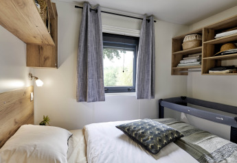 Bedroom in Mobile Home «New Horizon» at Flower Camping Provence Vallée, France, with bed, window, and shelves.