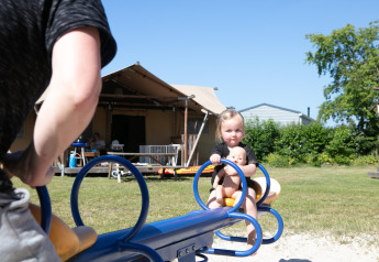 Bambina con bambola sull'altalena davanti a una tenda safari nel campeggio 't Veld, Paesi Bassi, giornata di sole.